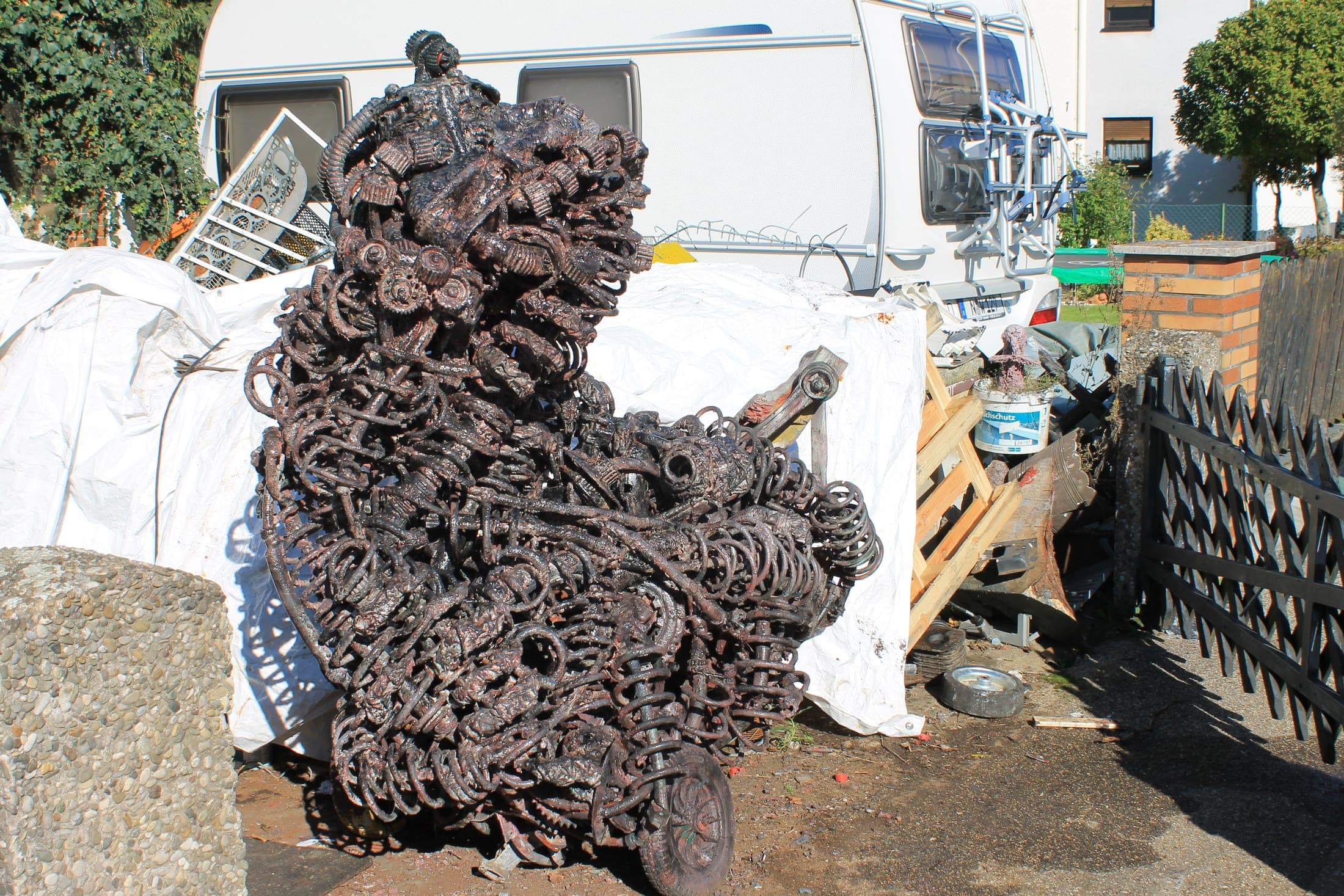 Aufrecht stehende, rostbraune Metallskulptur aus dicht verschweißten Ketten-, Rohr- und Federteilen.