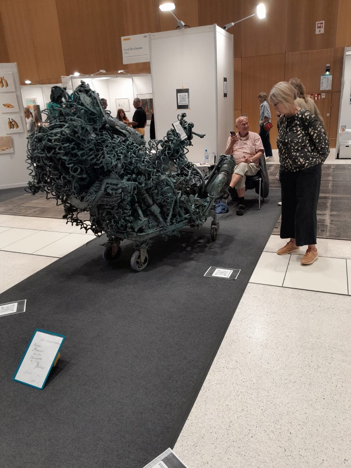 Lange, grün patinierte Metallskulptur aus unzähligen verschweißten Einzelteilen auf einem Rollgestell in einer Ausstellungshalle.