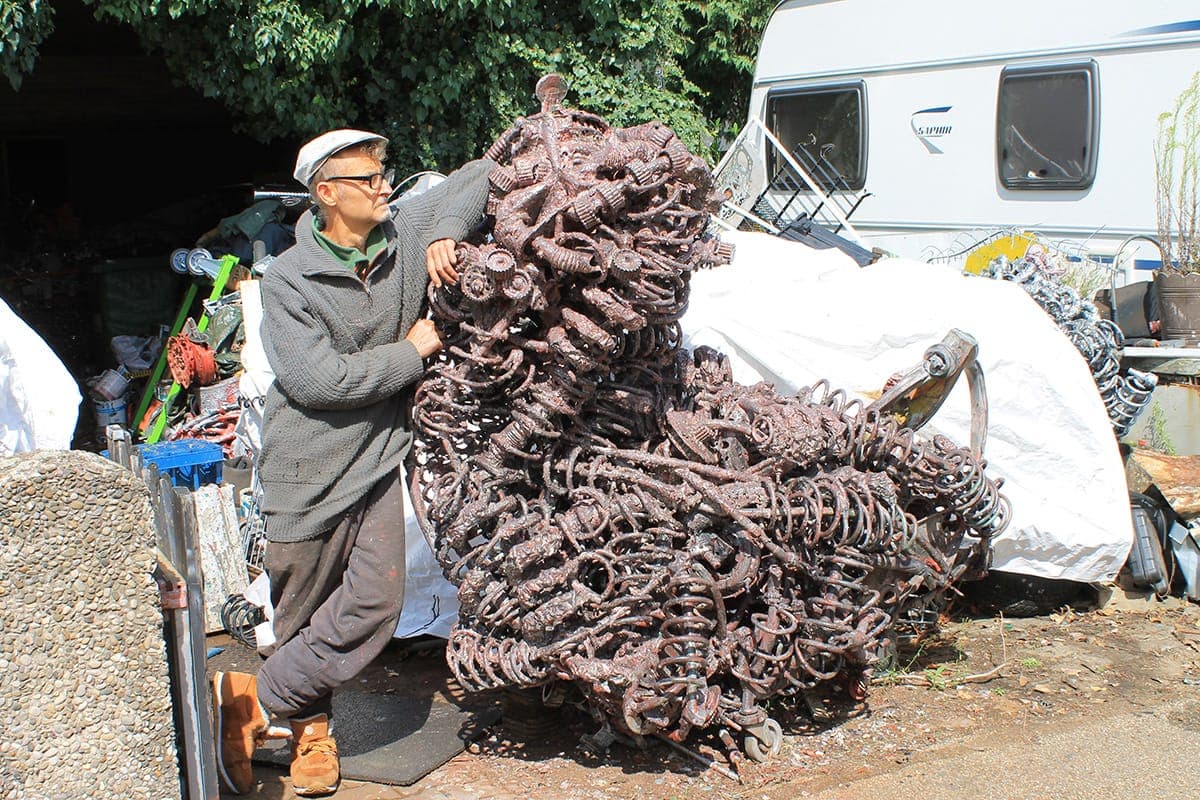 Gerd Georg Hirschmann steht mit Schiebermütze neben einer großen rostbraunen Metallskulptur im Hof seiner Werkstatt.