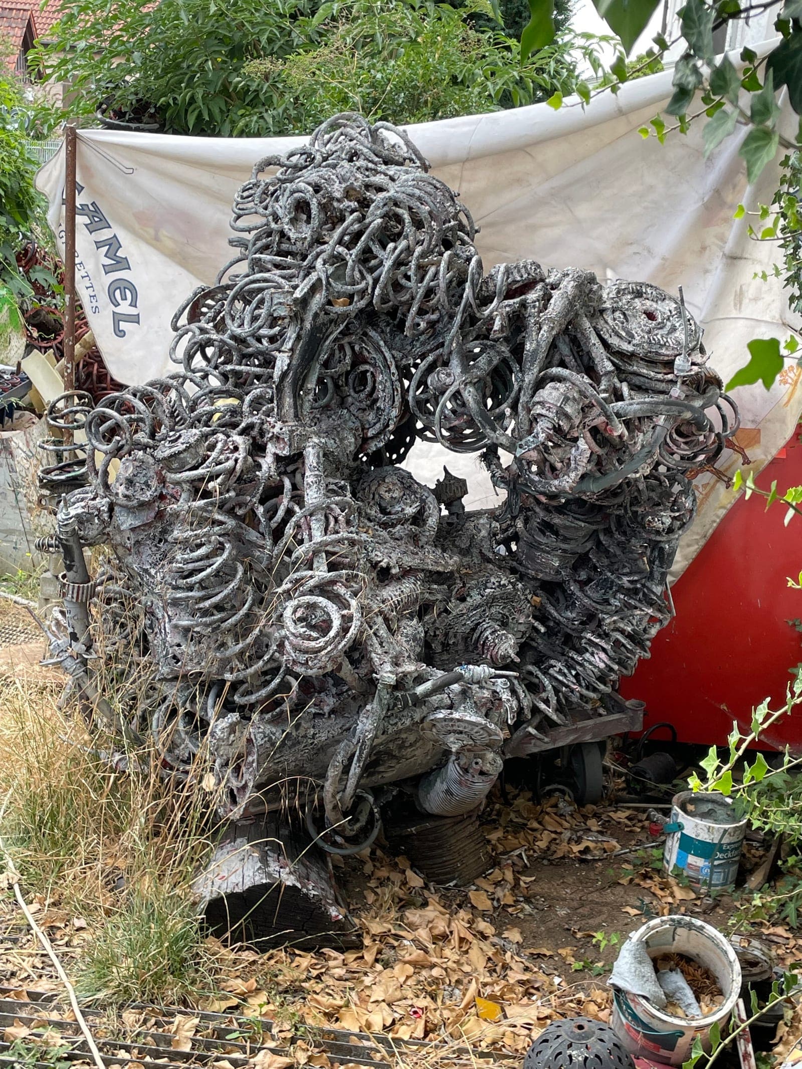 Hoch aufragende graue Metallskulptur vor einer hellen Plane, im Freien zwischen Werkstattgerät.