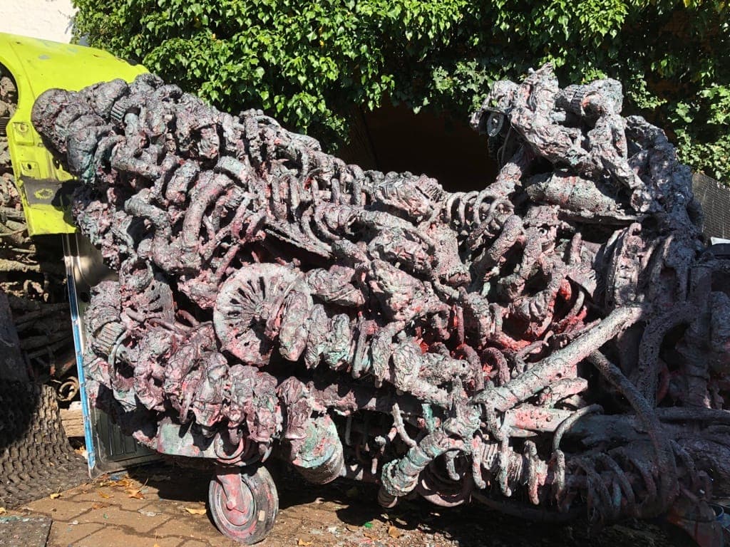 Mehrere Meter lange Metallskulptur auf Transportrollen, im Freien vor Grün und einer gelben Maschine.