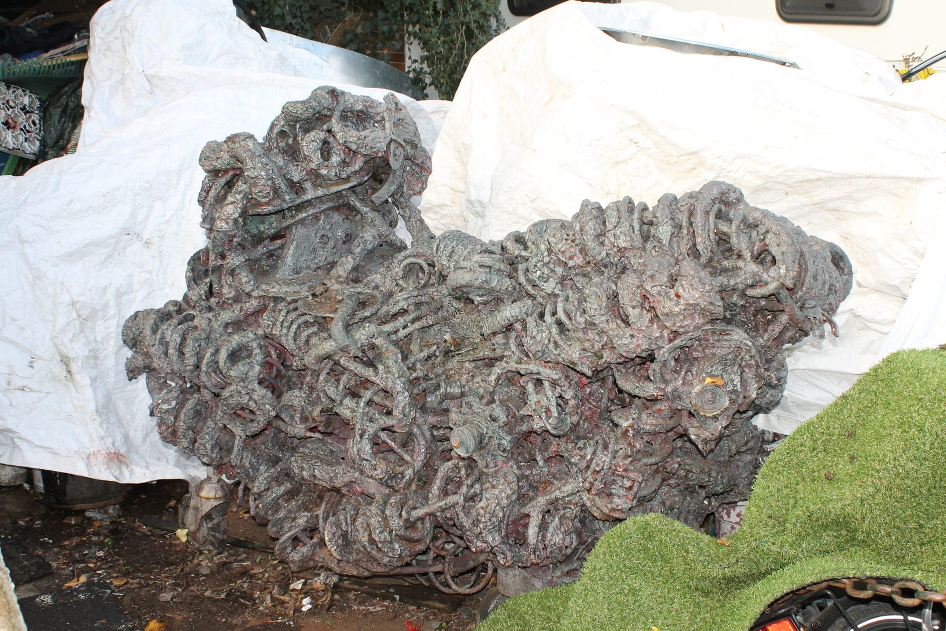 Breit gelagerte Metallskulptur mit rostroten und grauen Flächen, auf einer hellen Plane im Freien abgestellt.