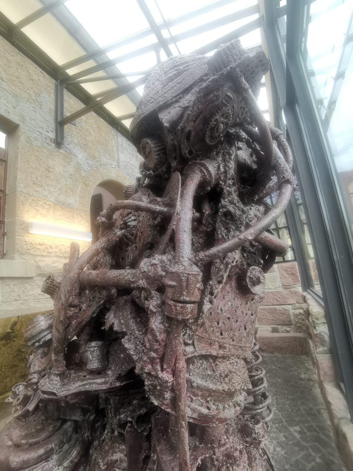 Dunkle, mannshohe Metallskulptur unter einem Glasdach vor einer hellen Natursteinwand.