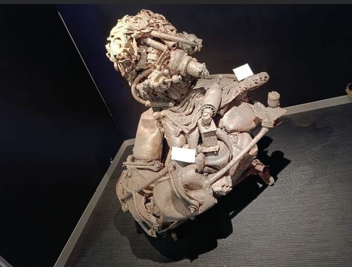 Kleinere Metallskulptur mit Werkschild auf einem Sockel in einem abgedunkelten Ausstellungsraum.