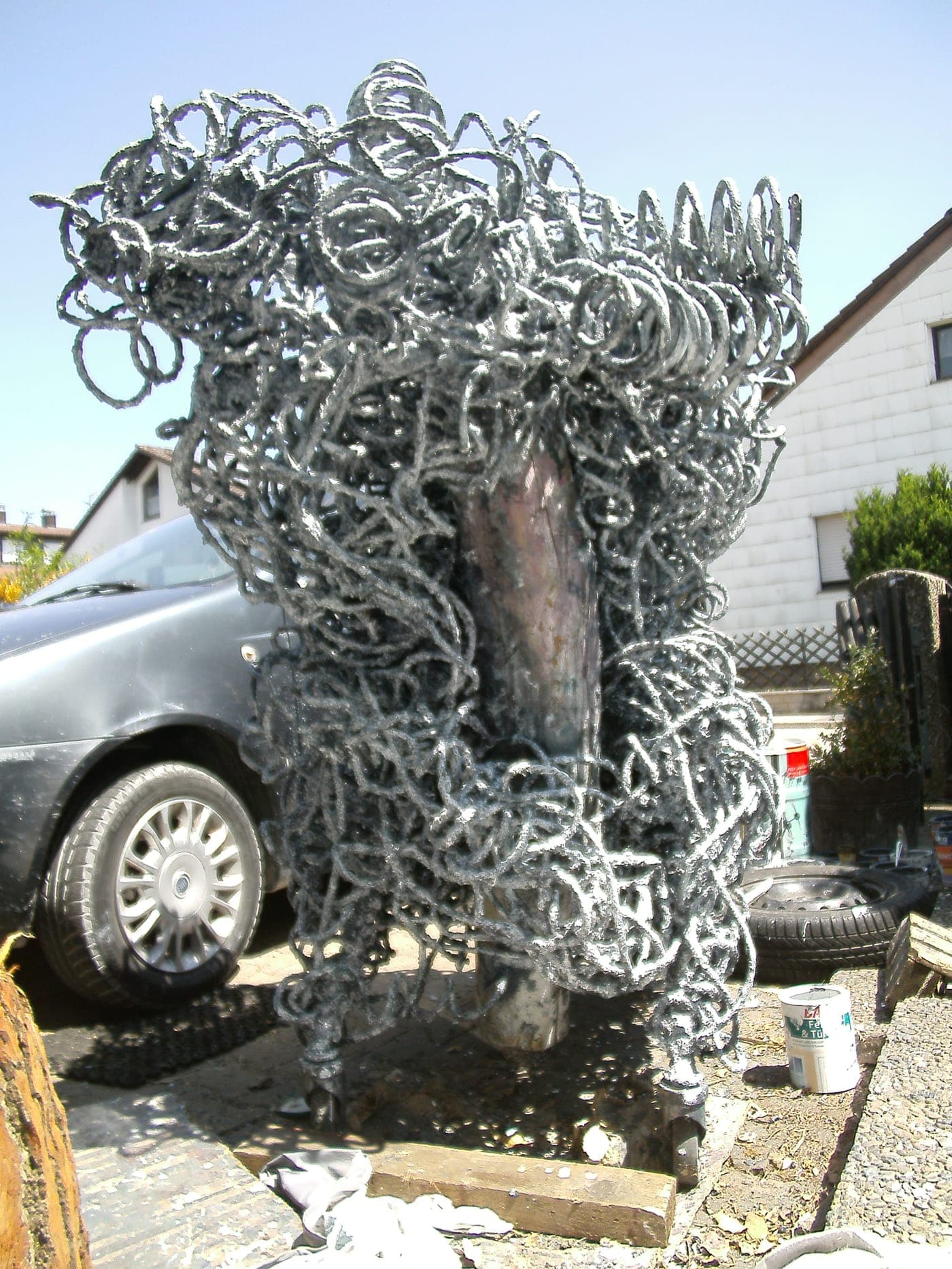 Silbrig glänzende Skulptur aus gewundenem Draht mit dunklem Kern, auf der Straße neben einem Fahrzeug abgestellt.
