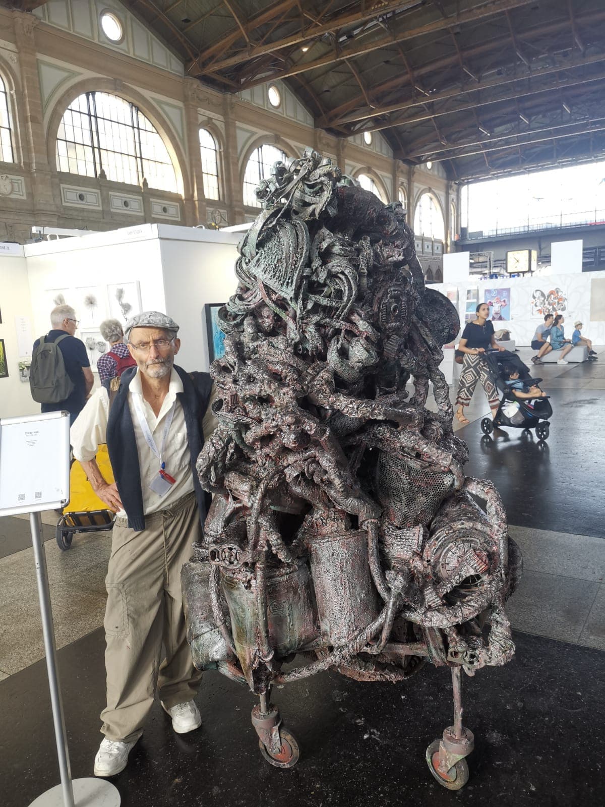 Der Künstler steht neben einer mannshohen Metallskulptur in einer historischen Bahnhofshalle mit Rundbogenfenstern.